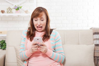 Şok olmuş, orta yaşlı, ağzı açık bir şekilde cep telefonuna bakıp evdeki kanepede oturan bir kadın. Boşluk ve yerel kavramı kopyala.