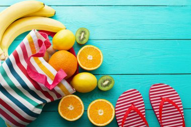 Yaz eğlencesi ve mavi ahşap arka planda meyveler. Model ve resim gibi. Portakal, limon, kivi, poşette muz meyvesi ve yerde parmak arası terlik. Üst görünüm ve kopyalama alanı