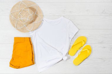 Beyaz yakın plan tişört düz yatar ve beyaz ahşap arka planda parmak arası terlikleri taklit eder. Üst görünüm ve boşluk kopyala. Yaz tişörtü ve yaz tatili maketi. Şablon boş gömlek.