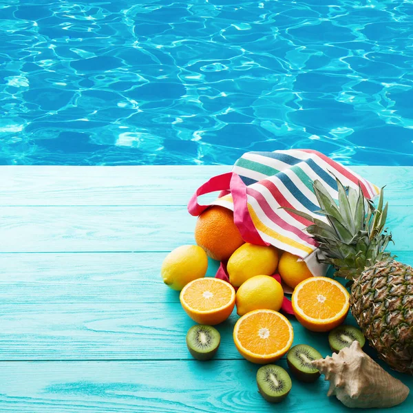 Citrus fresh food near swimming pool. Summer holidays. Orange, kiwi, pineapple, lemon and sea shell on blue wooden background. Vacation and summertime.Top view and copy space. Mock up