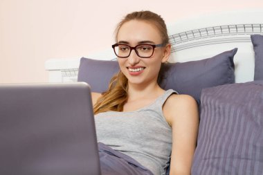 Sabah yatak odasında dizüstü bilgisayarının önüne uzanmış gözlüklü mutlu bir kadın.