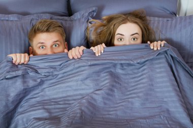 Bir adam ve bir kız yüzlerini bir battaniyenin arkasına saklıyorlar, mutlu bir çift yatakta.