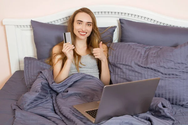Woman using laptop for store online shopping in bed at home, payment by credit card with online transaction banking. Girl holding creditcard and show like sign.