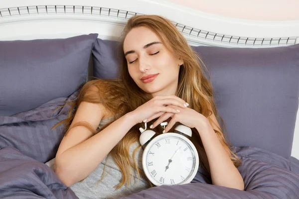 Concept of overslept work, bad sleep insomnia - Young beautiful sleepy girl with a closed eyes holds a clock and lies in her bed in the bedroom in the morning