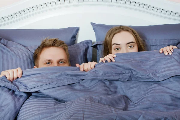 A guy and a girl hide their faces behind a blanket, a happy couple in bed
