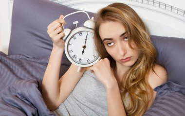 Genç, güzel, uykucu bir kız elinde bir saat tutuyor ve sabahları yatak odasında uzanıyor..