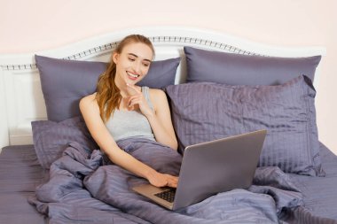 Güzel genç bir kadın laptopunu yatakta kullanıyor. Yatak odasında dizüstü bilgisayarda çalışan gözlüklü güzel kız.
