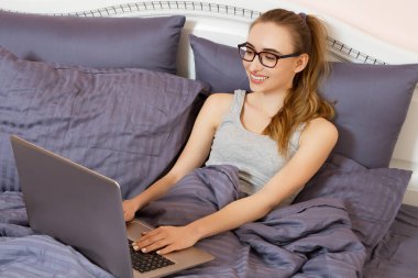 Güzel, gülümseyen beyaz bir kadının portresi dizüstü bilgisayarını kullanarak yatakta dinleniyor. Pozitif insan yüzü ifadesi müşteri memnuniyeti kavramı.