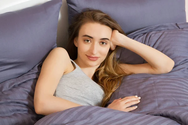 Yatağında uzanmış, gülümseyerek kameraya bakan bir kadın.