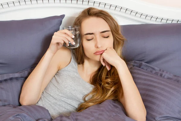 Elinde bir bardak su tutan ve yatakta yatan mutlu genç bir kadın. Kadın akşamdan kalma.