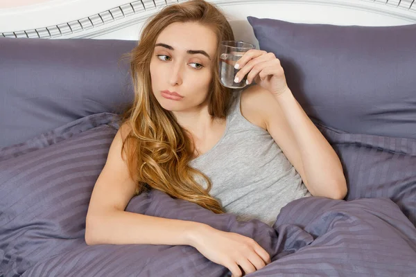 Güzel kız, elinde bir bardak suyla yatakta yatıyor. Akşamdan kalma ve başı ağrıyor..