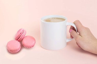Beyaz kupa modeli. Pembe badem kurabiyesi ve sütlü kahve. Şeftali renkli arka planda bir kadının el ele tutuşması. Tasarım tanıtımı için model