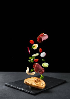 Siyah arka planda bir sandviç. Malzemeler havada uçuşuyor. Metin için boş alan. Veteriner ve sebzeli sandviç..