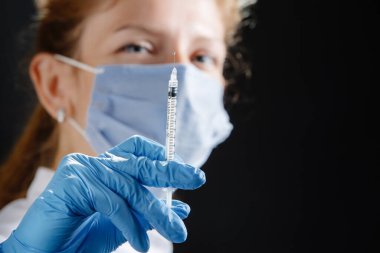 Elinde şırıngayla kameraya bakan genç bir kadın doktor. Tıbbi maske ve mavi lateks eldiven takan bir hemşire. İlaç ve sağlık hizmetleri. Virüs aşısı için. Önleyici tedbirler