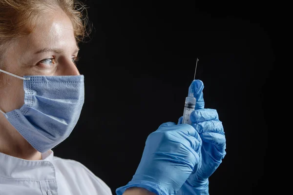A female doctor wearing a protective mask and gloves holds a syringe with a vaccine or injection against a black background. A female nurse prepares to give an injection against a dark background