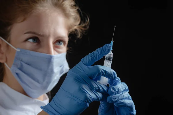 A female doctor wearing a protective mask and gloves holds a syringe with a vaccine or injection against a black background. A female nurse prepares to give an injection against a dark background