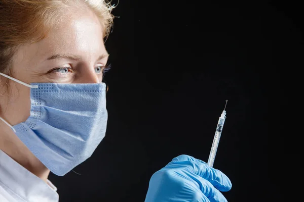 Portrait of a doctor or nurse in a medical uniform and protective blue mask with a syringe in his hand on a black background.