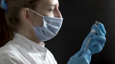 A middle-aged woman doctor wearing a protective mask and protective gloves on a black background draws a syringe of medicine, a vaccine, a shot. The nurse makes preparations for the vaccination or