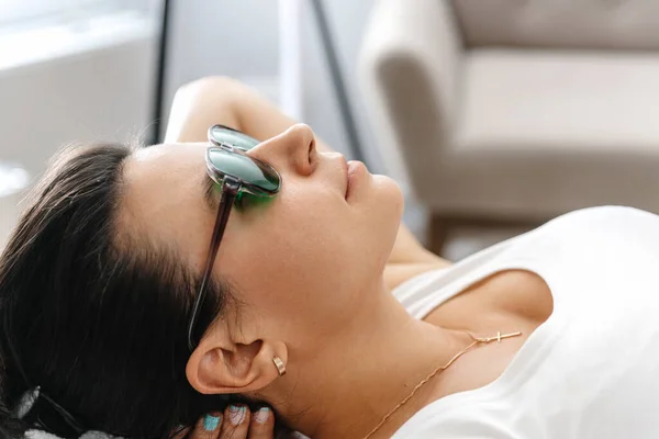 a girl in protective goggles for laser hair removal lies on her back during the procedure. A cosmetologist performs laser facial treatment for a woman at a beauty spa. Laser facial hair removal