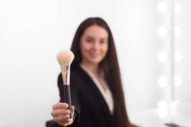 Kız, makyaj sanatçısı, iş elbisesi içinde uzun siyah saçlı uzanmış bir elle toz fırçası tutuyor, beyaz arka planda.