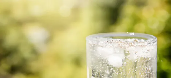 Glas drinkwater met bubbels, ijsblokjes om het koud te maken, natuurlijke wazige achtergrond met lichten en schaduwen. concept van zuiverheid, drinkwater, gezonde levensstijl of medische — Stockfoto