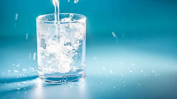 Vaso de agua potable con burbujas, agua está vertiendo, fondo azul estudio con luces y sombras. concepto de pureza, agua potable, estilo de vida saludable o medicina — Foto de Stock