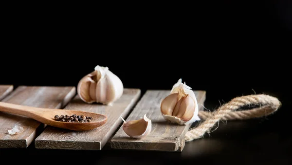 Fresh organic garlic over black background, full heads, cloves and peels, wooden spoon with black pepper, cooking ingredients — Stock Photo, Image
