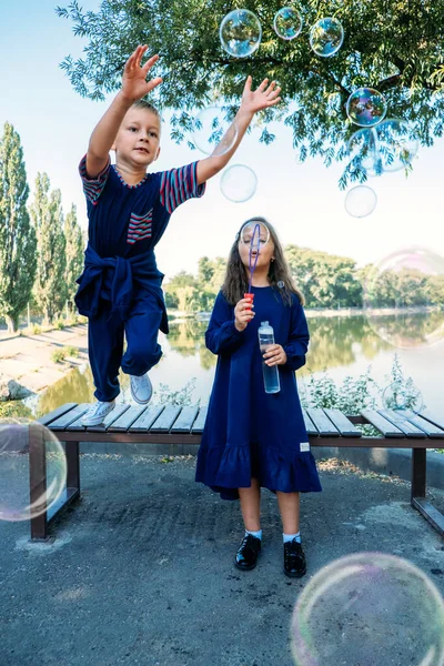 Glückliche Jugendfreundschaft. Kleines Mädchen pustet Seifenblasen und aufgeregter Junge springt und fängt Seifenblasen. Glückliche Kinder Bruder und Schwester genießen Seifenblasen machen. — Stockfoto