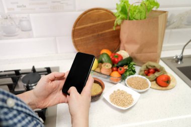 Arka plandaki mutfak masasında boş ekranlı cep telefonu, sebzeler, meyveler, yemek malzemeleri. Çevrimiçi yemek konsepti için akıllı telefon modeli