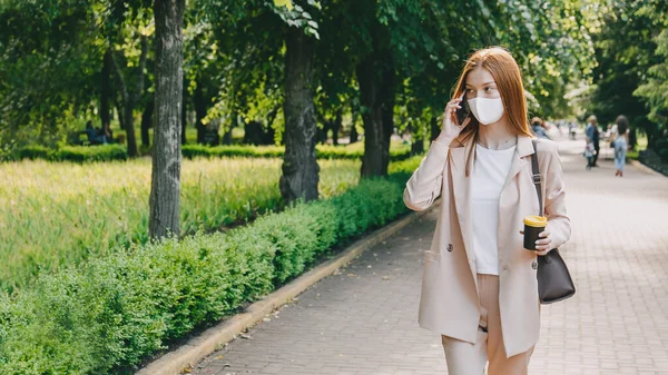 Donna rossa che indossa una maschera per la protezione dai virus. Giovane donna d'affari con il cellulare in mano passeggiando nel verde del parco estivo. — Foto Stock