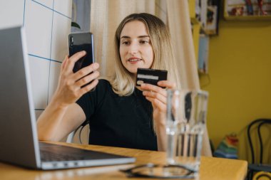 İnternet Satın Alma, Çevrimiçi Alışveriş, Alışveriş, E-ticaret sitesi, akıllı telefon ya da dizüstü bilgisayar. Evde elinde kredi kartı olan, dizüstü bilgisayar ve cep telefonu kullanan genç bir kadın.