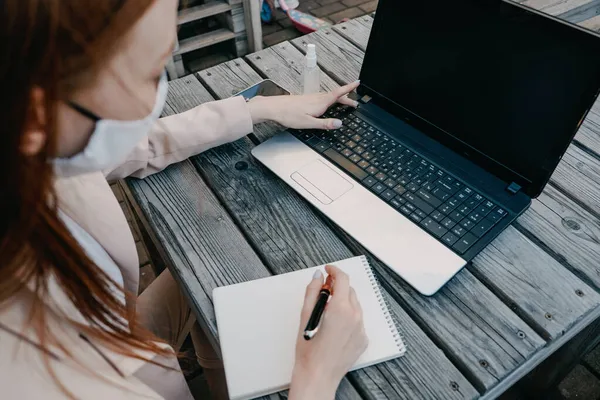 Pelirrojas Mujer de negocios con máscara facial para la protección contra el virus que trabaja en el ordenador portátil al aire libre. Joven empresaria trabajando en el portátil fuera. — Foto de Stock