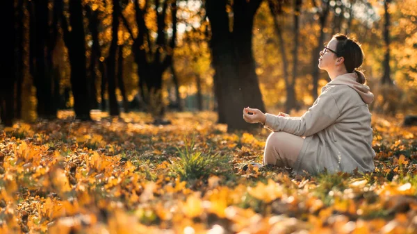 Mulher de meia-idade branca meditando em pose de lótus no parque de outono com luz solar. Yoga no parque de outono. Mulher de óculos relaxante em folhas amarelas. Meditação, Saúde mental, autocuidado, atenção plena — Fotografia de Stock