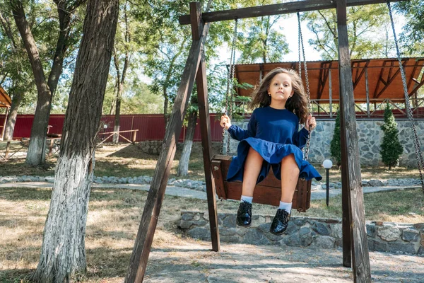 Szczęśliwa dziewczynka w niebieskiej sukience na huśtawce w letnim parku. Słodka dziewczyna w parku huśtawka na huśtawce. Letni aktywny wypoczynek i aktywność na świeżym powietrzu — Zdjęcie stockowe