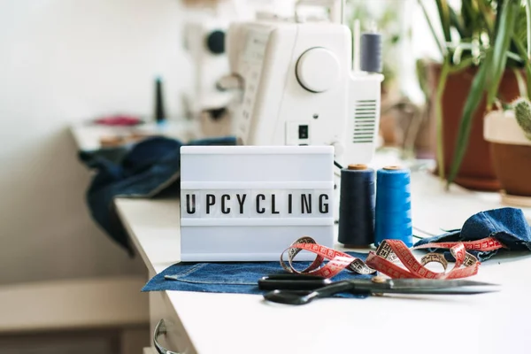 Upcycling text on light board on sewing machines with old blue jeans background. Denim clothes, scissors, thread and sewing tools in sewing studio. Denim Upcycling and repair — Fotografia de Stock