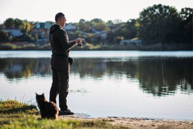 Çevresindeki doğa arka planında olta ve kedi taşıyan balıkçı. Balık tutan balıkçı ya da nehir kenarındaki olta kamışı. Çubuklu balıkçı, nehir kıyısında dönen bobin.