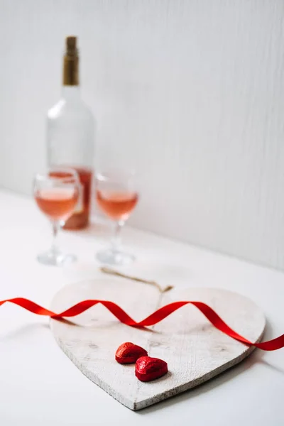Праздничный стол с двумя бокалами розового вина, шоколадными конфетками в форме красного сердца и красными лентами на белом фоне. День святого Валентина, романтика, концепция знакомств, пространство для копирования — стоковое фото