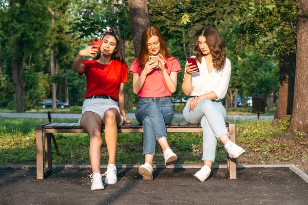 Diversas tres amigas usando sus teléfonos al aire libre. Grupo de personas jóvenes gen z utilizando teléfono inteligente móvil sentado en el banco en el parque de verano — Foto de Stock