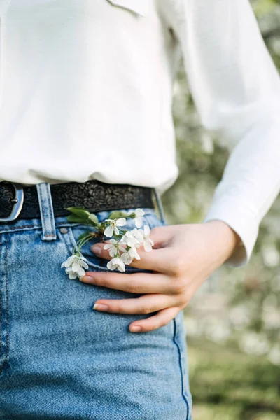 A primavera está a chegar. Beleza natural. Mão feminina no bolso de calça jeans jeans com flores brancas no jardim de primavera. Mão de mulher e flores de cereja branca em jeans jeans jeans azul — Fotografia de Stock