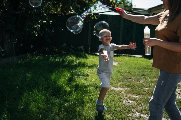 Glad familj blåser såpbubblor i trädgården, trädgård. Barnunge pojke blåser såpbubblor. Lycklig ung mor och hennes son blåser såpbubblor i parken — Stockfoto