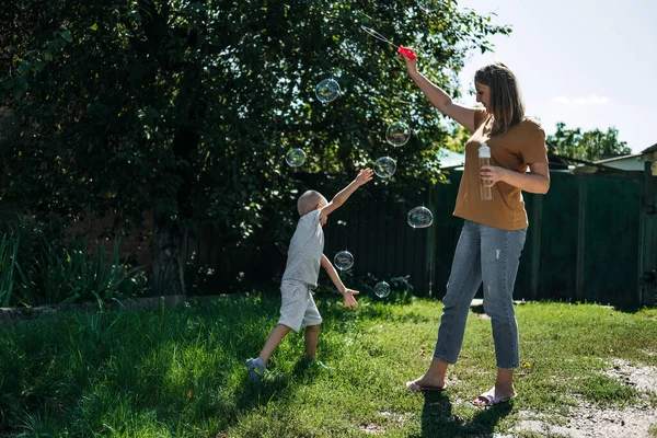 Happy family blowing soap bubbles in the backyard, garden. Child kid boy blowing soap bubbles. Happy young mother and her son blowing soap bubbles in park — Zdjęcie stockowe