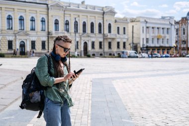 City Summer Vacation, urban trip. Exploring new places, cities around world. City travel outfit. Hipster woman traveler with dreadlocks headphones and cell phone on summer city background