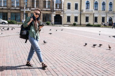 City Summer Vacation, urban trip. Exploring new places, cities around world. City travel outfit. Hipster woman traveler with dreadlocks headphones and cell phone on summer city background