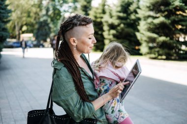 Serbest ebeveynler. Anne ve bebek kızıyla el üstünde yürüyüp sokakta tablete bakmakla meşguldüm. Bağımsız anne bebeğini kucağında tutarken notlar alıyor..
