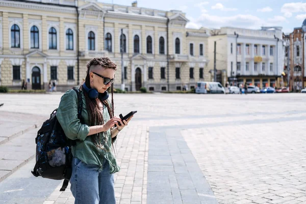 Şehir Yaz Tatili, şehir gezisi. Yeni yerler, dünyadaki şehirleri keşfetmek. Şehir seyahati kıyafeti. Hipster kadın gezgin rastalı kulaklıklı ve cep telefonlu yaz şehri arka planında