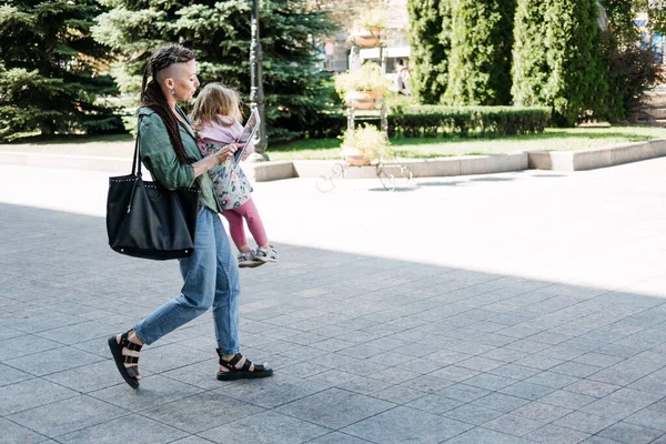 Freelance Parents. Busy working mom with baby toddler daughter on hands walk and looking on tablet in street. Multitasking Freelancer mom making notes while holding her baby outddors. — стоковое фото