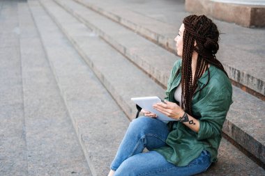 Serbest meslek, yan iş. Sokakta tabletle çalışan dövmeli ve rastalı genç bir hippi kadın. 30 'ların moda kadını açık hava tabletine bakıyor..