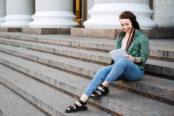 Freelance, side hustle. Young woman hipster freelancer with tattoo and dreadlocks working with tablet on the steps of the street. 30s trendy woman is looking into her electronic tablet outdoor. — стоковое фото