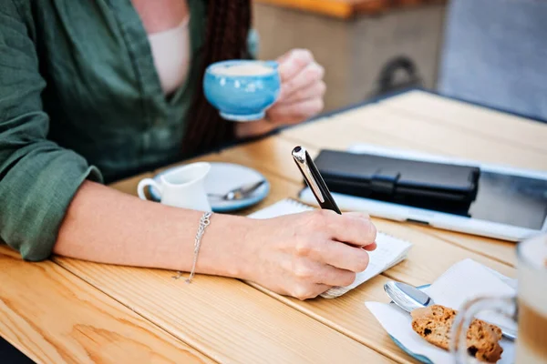 To do list, planning for freelancers. Woman holding pens and papers making notes and drinking coffee in street cafe. Close-up of female freelancer writing and drawing in notebook —  Fotos de Stock