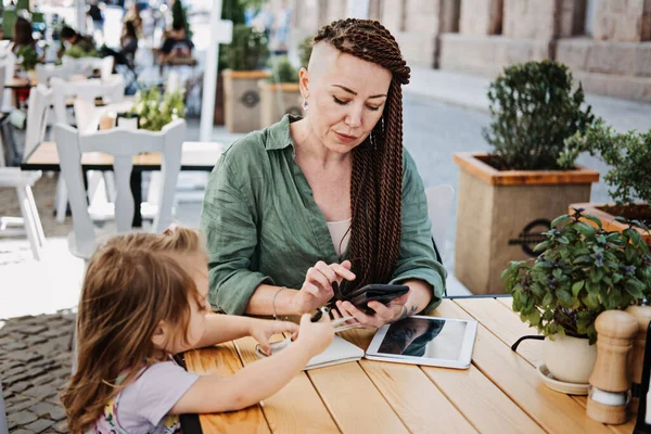 Freelance Parents. Busy working mom with cell phone and tablet working in street cafe with baby toddler daughter. Multitasking Freelancer mom and her baby daughter working together outdoors. — Stock Photo, Image